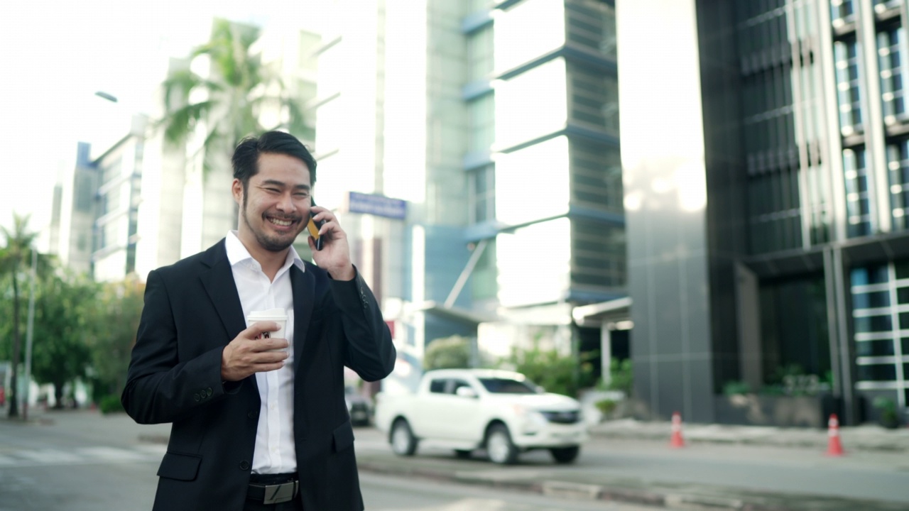 亚洲商人穿着黑色西装微笑着走在城市的街道上上班。手拿咖啡杯使用智能手机。城市lifeStlye概念。视频素材