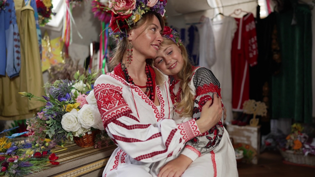 快乐的母女穿着乌克兰民族服装，坐在室内拥抱着，望向别处。美丽的女人和漂亮的女孩的肖像在工艺商店在新的裙子。缓慢的运动。视频素材
