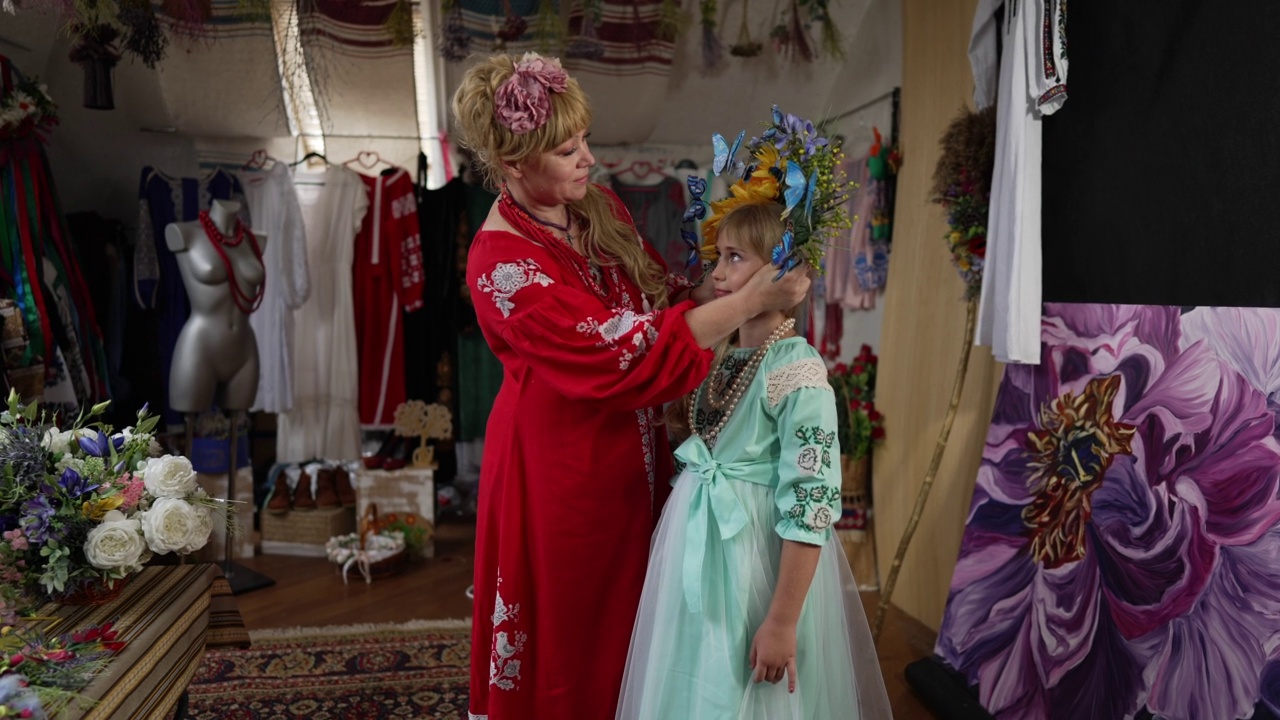 快乐的乌克兰母亲戴上传统的花环，可爱的女儿用慢动作说话。积极的美丽的女人和漂亮的女孩的肖像站在室内的民族服装。视频素材
