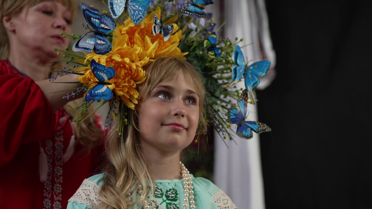 可爱的乌克兰小女孩的特写肖像在美丽的头花环与模糊的女人调整配件在慢动作。微笑迷人的漂亮女儿和母亲在室内。视频素材