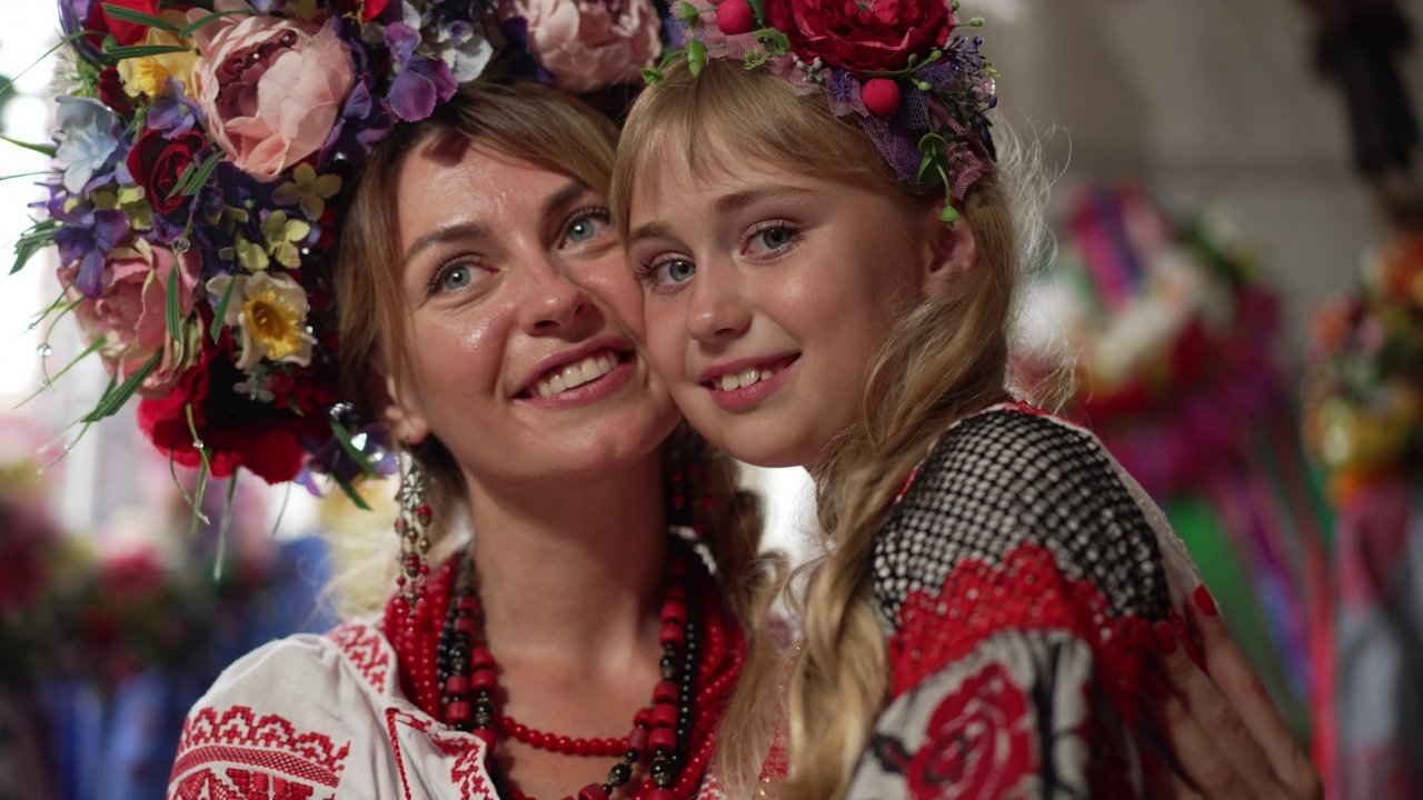 近距离迷人的女孩微笑着看着相机，美丽的女人拥抱孩子。可爱的乌克兰女儿穿着民族服装，头戴花环，和母亲在室内用慢镜头摆姿势。视频素材