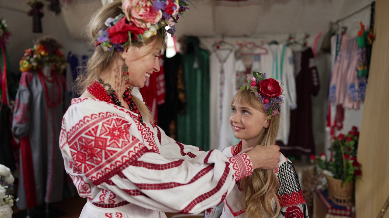 微笑的乌克兰妇女爱抚着长头发的快乐女孩在传统的刺绣服装。美丽的年轻母亲和迷人的女儿在室内慢动作交谈。家庭的概念。视频素材