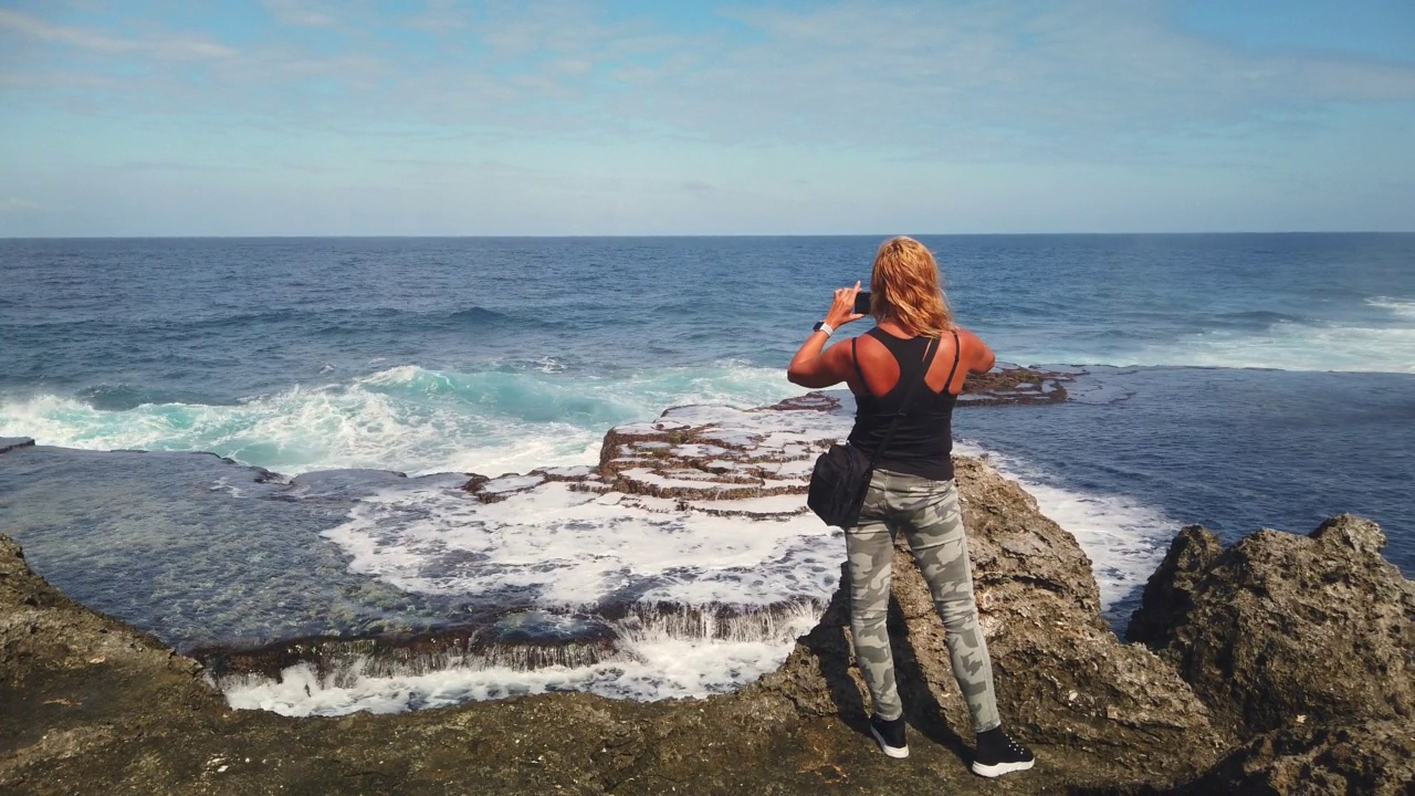 图为，在暴风雨来临之前，女游客正在用智能手机拍摄海浪翻滚的悬崖的照片或视频。视频下载