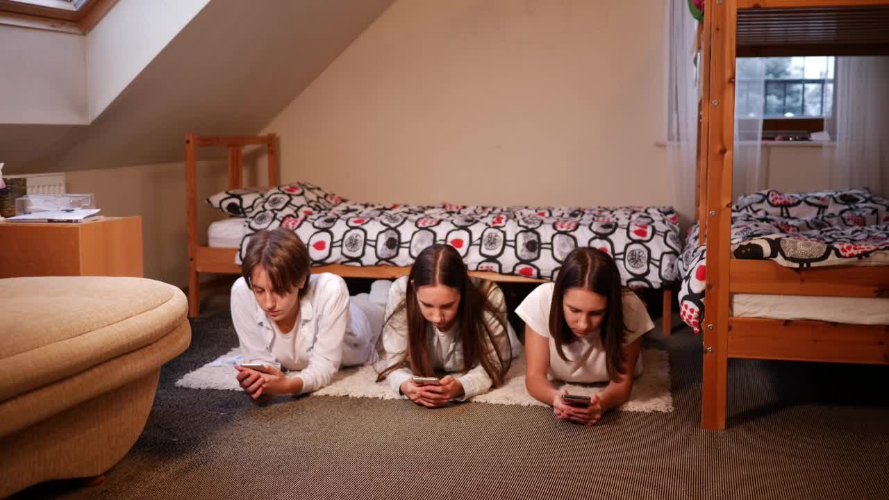 三姐妹十几岁的女孩在家里使用智能手机，躺在卧室的地板上，花时间在社交媒体视频下载