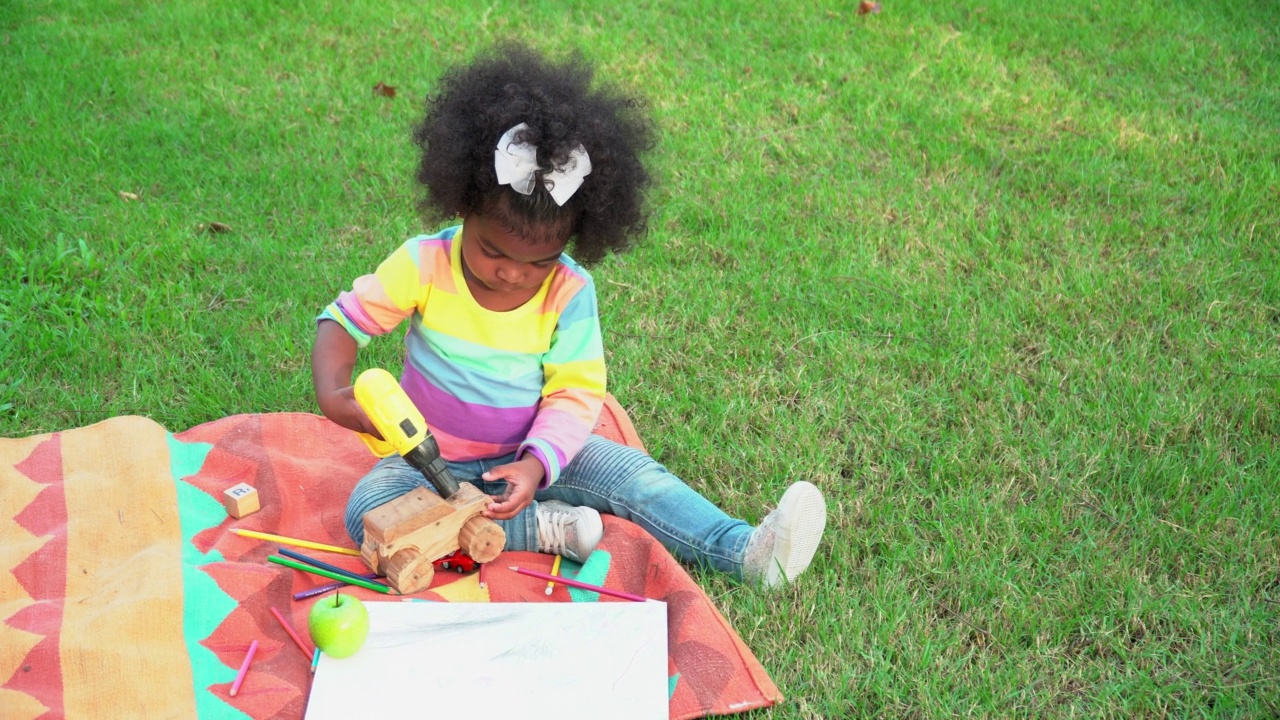 一个可爱的美国非洲小女孩穿着彩色的布和牛仔裤和大的发弓坐在野餐毯子在绿色的公园里玩玩具，汽车和枪。的童年。感受快乐，户外教育理念视频下载