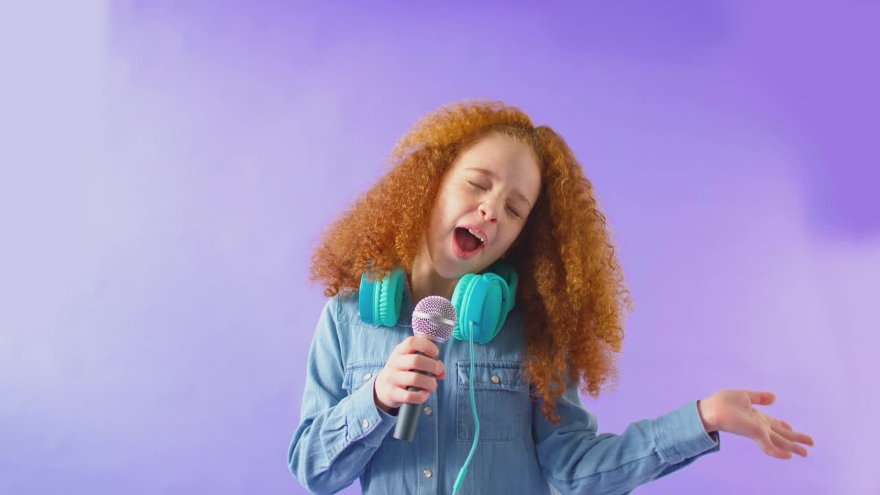 演播室拍摄的女孩戴着耳机唱卡拉ok进入麦克风对紫色的背景-拍摄在慢动作视频下载