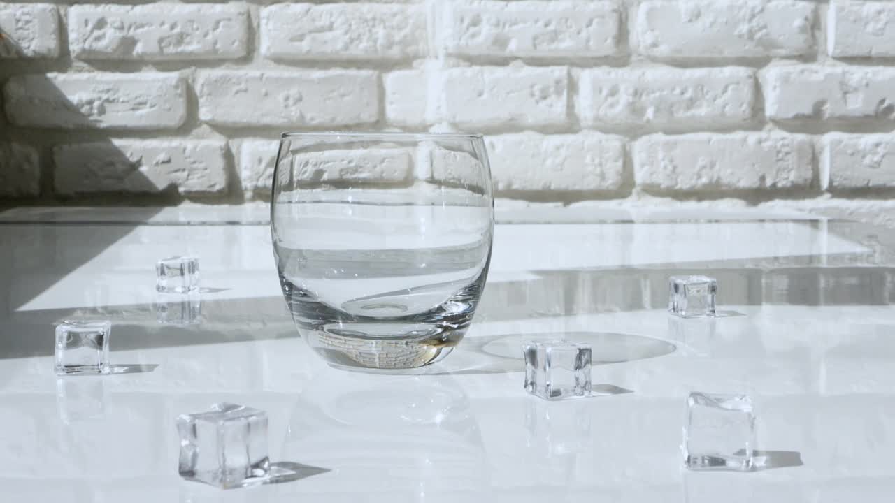 将水倒入一个透明的玻璃杯中，在白色砖墙的背景上有一个冷冻矿泉水饮料。视频素材