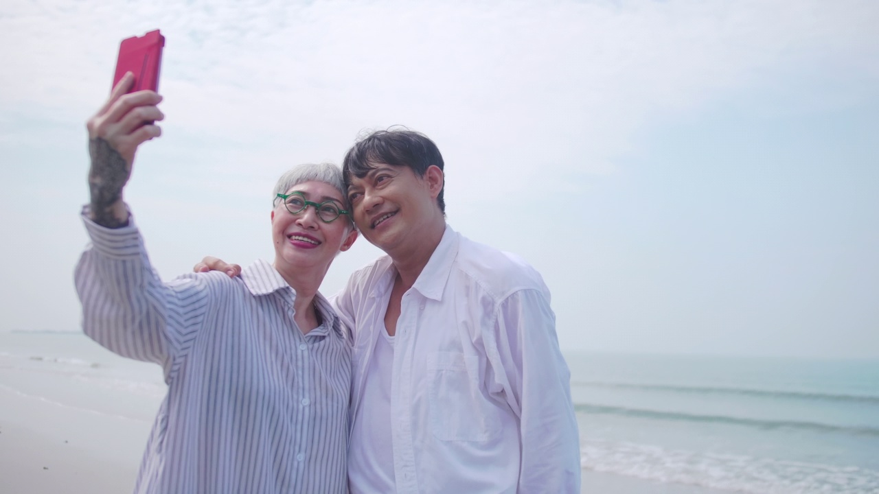 亚洲退休夫妇在夏天的海边放松。一名老年女性在沙滩上和她的伴侣自拍。视频下载