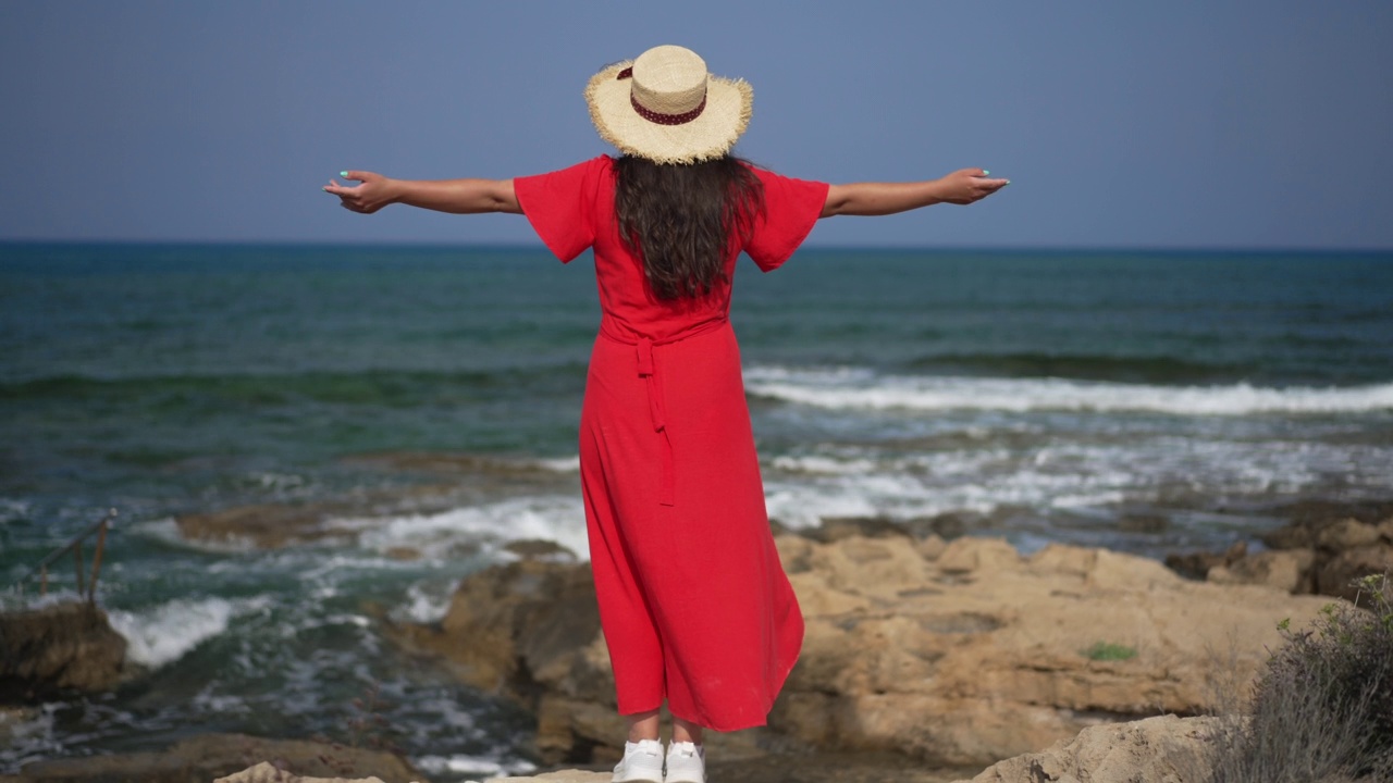 后视图年轻兴奋的女人在红色连衣裙站在地中海海滩举起手在慢动作。广角镜头难以辨认的快乐高加索游客欣赏自然享受假期。视频素材