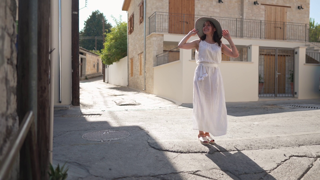 宽镜头肖像的年轻快乐的女人在白色连衣裙站在塞浦路斯的阳光街道上举起手在慢动作。带着草帽微笑着欣赏古镇的美丽白人游客。视频素材