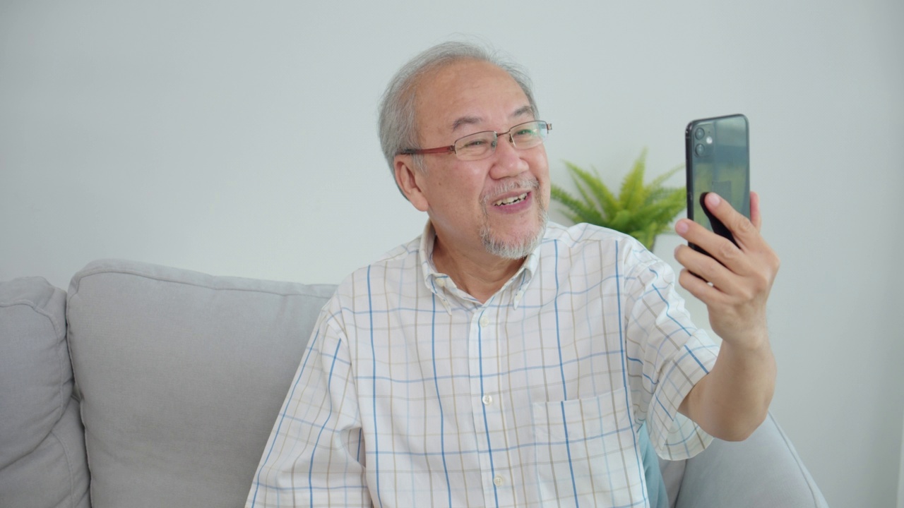 亚洲老人愉快地拿着手机进行在线视频通话，说话，微笑和大笑。老人积极学习使用现代科技设备，在家里探索小玩意的功能。视频下载