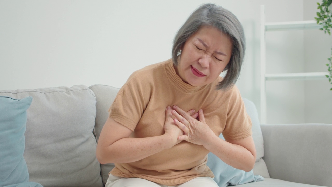 一位年长的亚洲妇女坐在沙发上，双手放在胸前，因为她感到胸闷，胸口有灼痛，在她家的客厅里。视频下载
