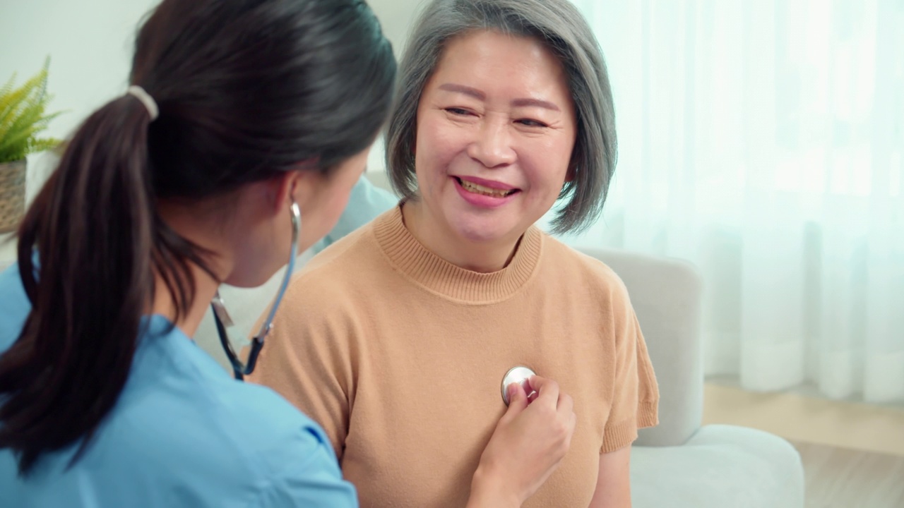 亚洲女医生使用听诊器检查一位年老的亚洲女病人的呼吸和心跳。老妇人坐在沙发上微笑着和医生畅谈。视频下载