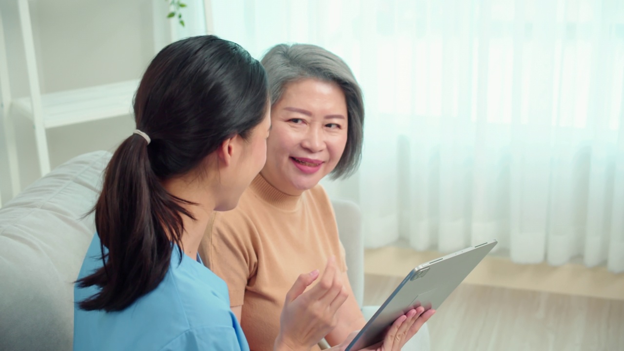 亚裔女性专业护理人员检查并出示体检结果，解释处方，使用药片访问坐在家里客厅沙发上的亚裔老年女性患者。视频下载