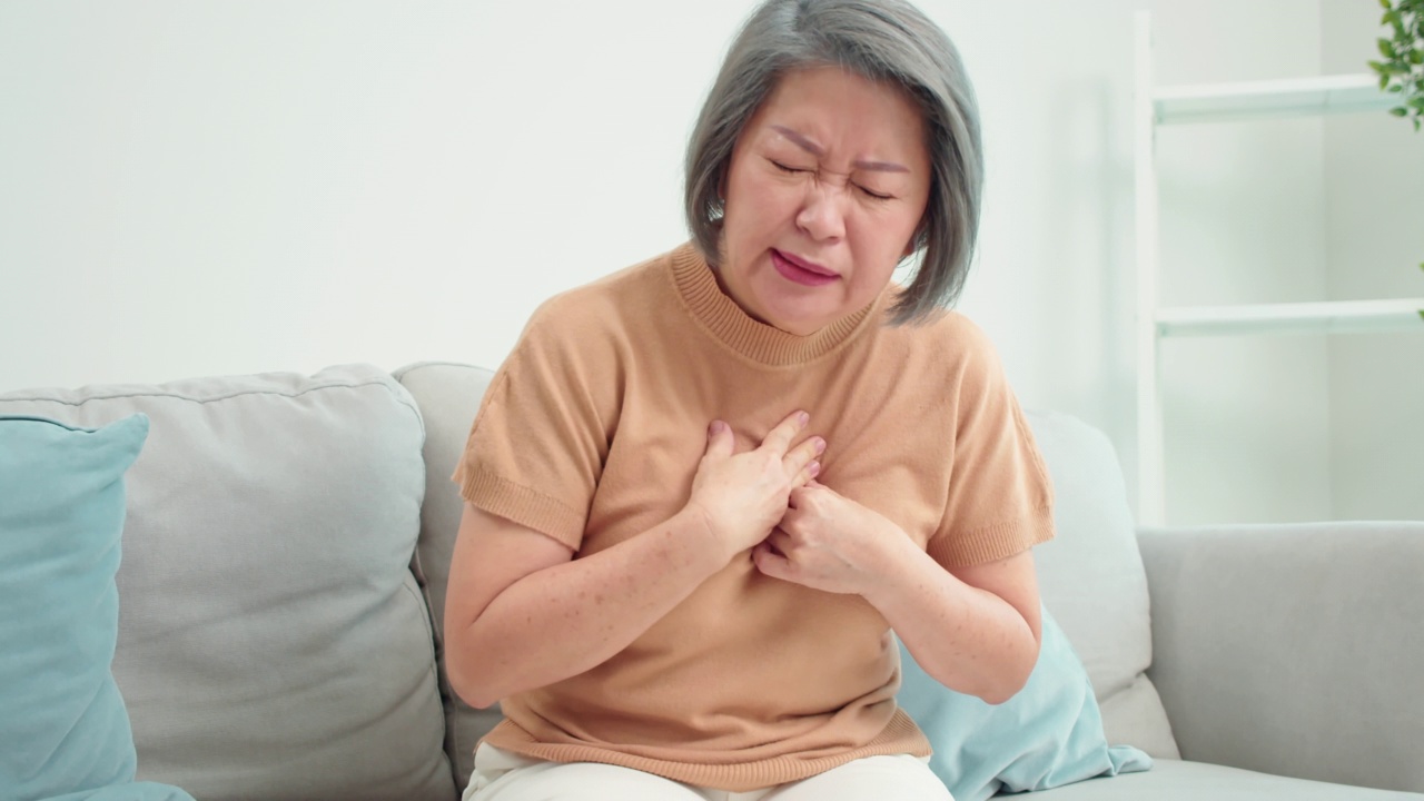 一名亚洲老年妇女坐在家里客厅的沙发上心脏病发作。患有痛苦和胸痛的老年女性因剧烈疼痛而呼吸困难。视频下载