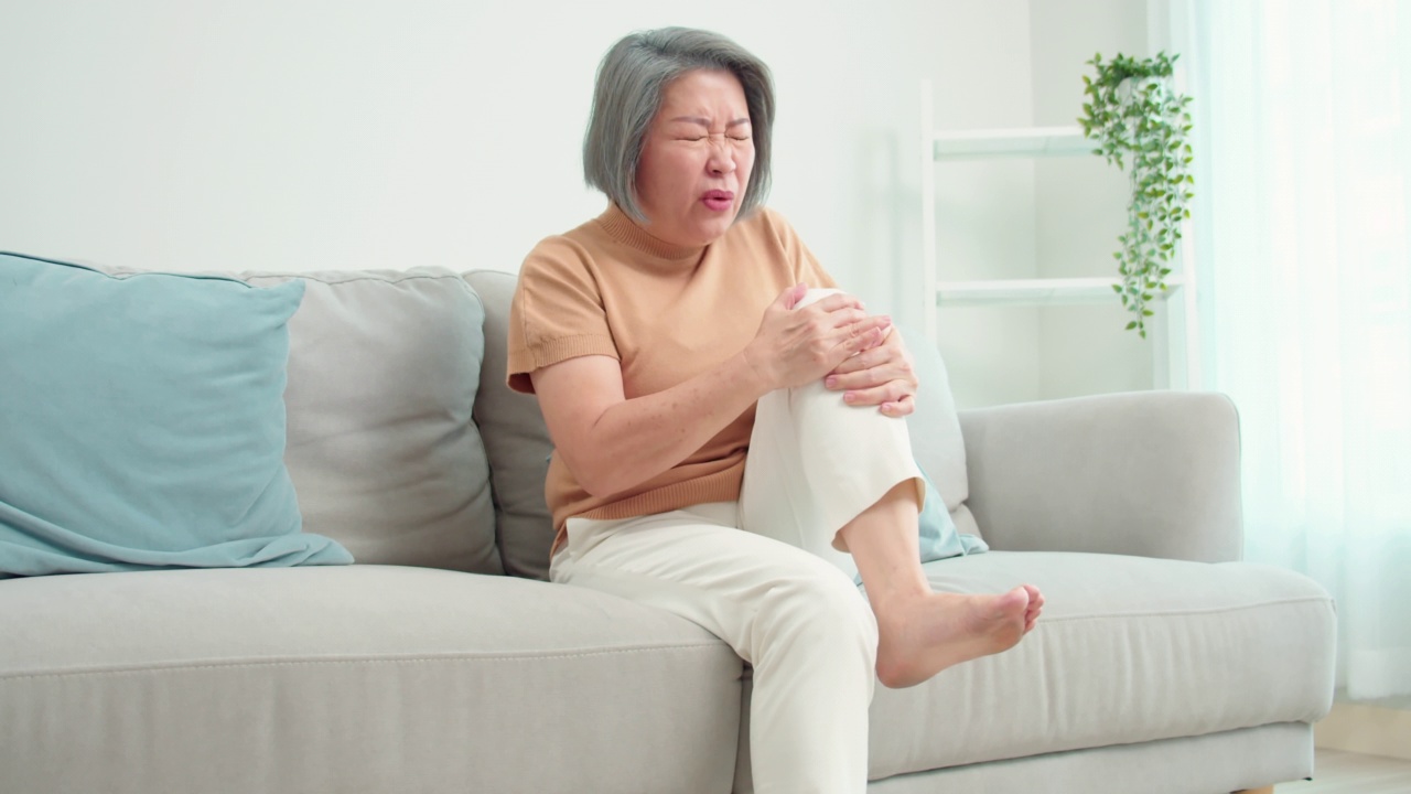 图为，从客厅的沙发上起身时膝盖疼痛的亚洲老年女性的特写。患有骨关节炎的老年女性。健康问题与人的观念。视频下载