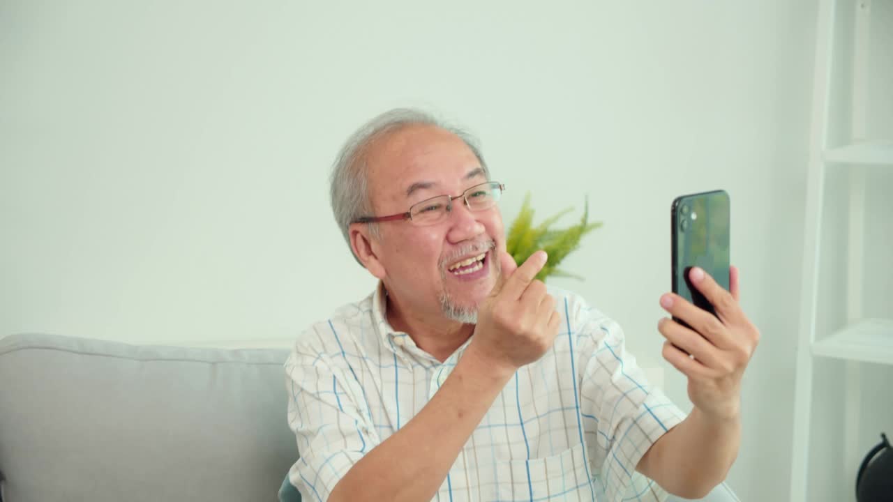 亚洲老人愉快地拿着手机进行在线视频通话，说话，微笑和大笑。老人积极学习使用现代科技设备，在家里探索小玩意的功能。视频下载