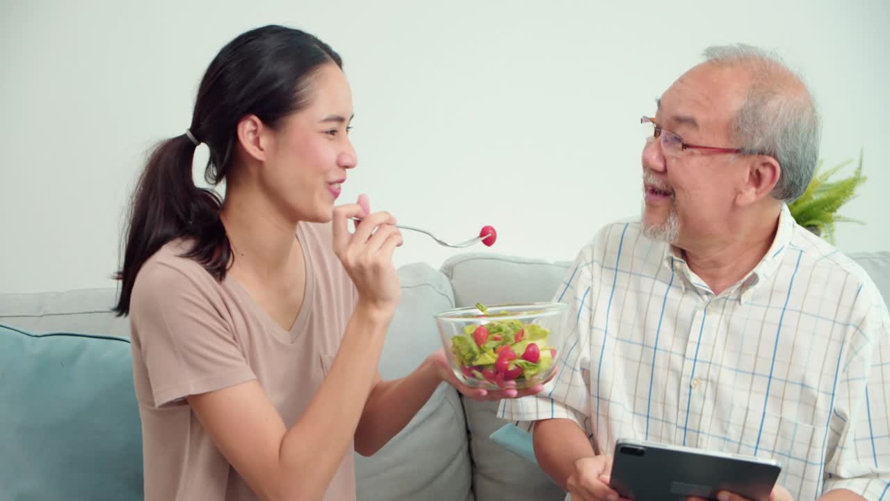 照顾年轻的亚洲妇女喂蔬菜，年长的亚洲男子坐在沙发上的客厅在家里。女性治疗师正在监测老年男性患者的症状。医疗保健医疗的概念。视频下载