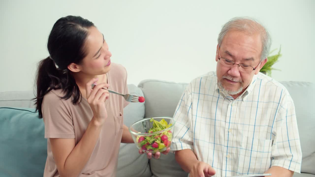 照顾年轻的亚洲妇女喂蔬菜，年长的亚洲男子坐在沙发上的客厅在家里。女性治疗师正在监测老年男性患者的症状。医疗保健医疗的概念。视频下载