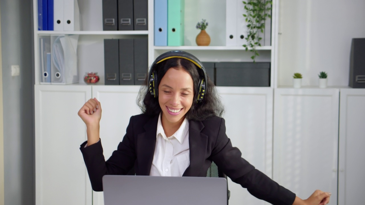 美丽的拉丁商业女性坐在和戴耳机在办公桌在办公室工作场所。快乐和微笑的女人听音乐放松，跳舞和享受自己。工作中放松的概念。视频下载