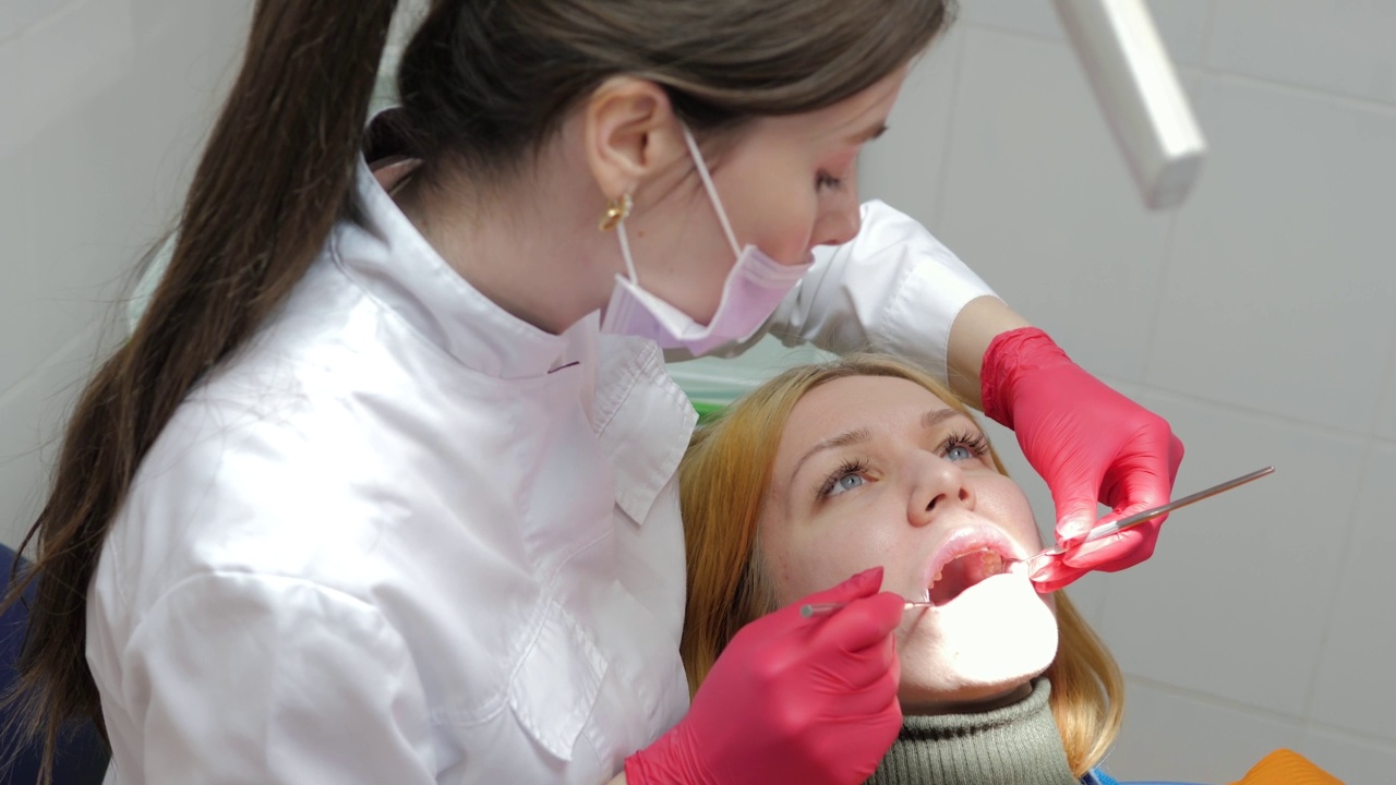 一个戴着面具的牙医在牙医预约时检查一个迷人的女孩的牙齿。戴着手套的牙医在病人的口腔内操作牙科器械。牙科诊所。牙科治疗。视频素材