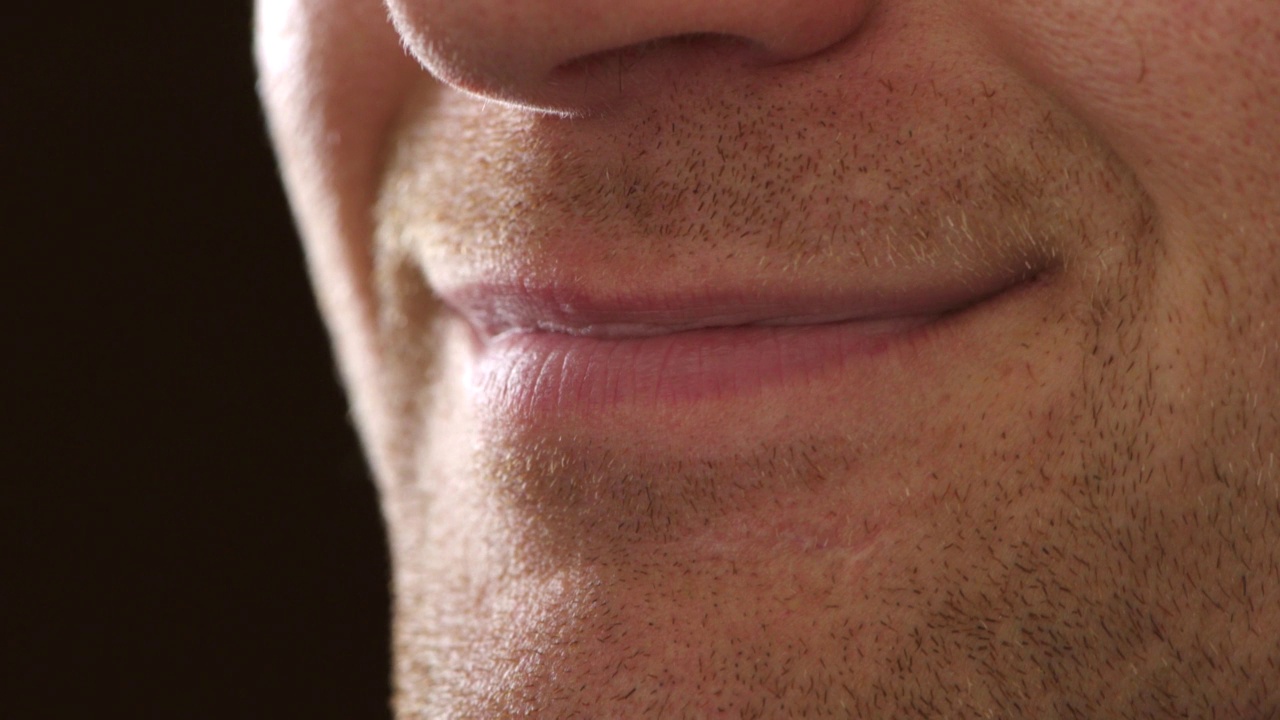 一个害羞的男人的嘴，给一个紧张或不确定的微笑，感到焦虑或不舒服在演播室的黑色背景。特写的嘴唇和牙齿的男性胡子茬微笑视频素材