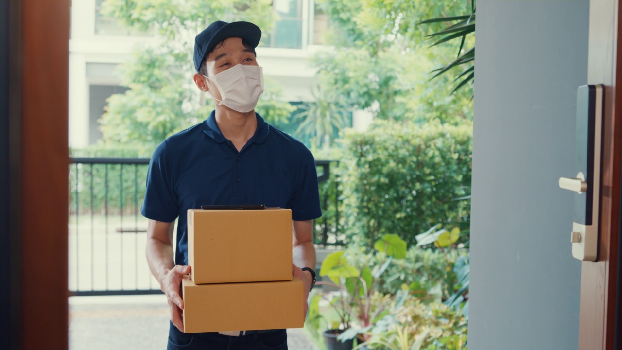 年輕的亞洲快遞員戴著面具，穿著藍色海軍制服攜帶箱子箱子敲門回家網上購物紙箱給女人在門口女士掃描二維碼支付在家里的智能手機。視頻下載