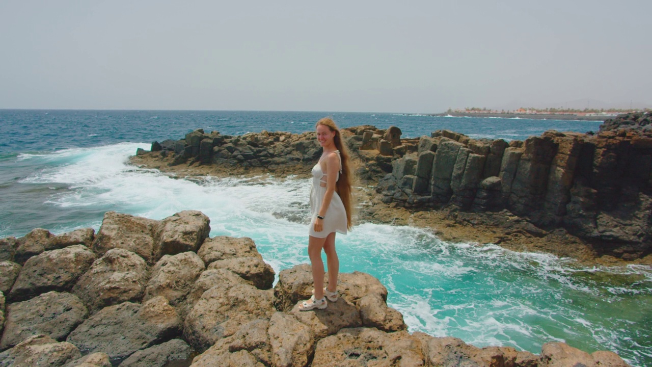 Fuerteventura,加那利群岛。迷人的多种族女子享受天堂，阳光明媚的日子在清澈湛蓝的海洋岩石海边放松，风吹头发。海浪拍打着火山峭壁，浪花翻滚。视频下载