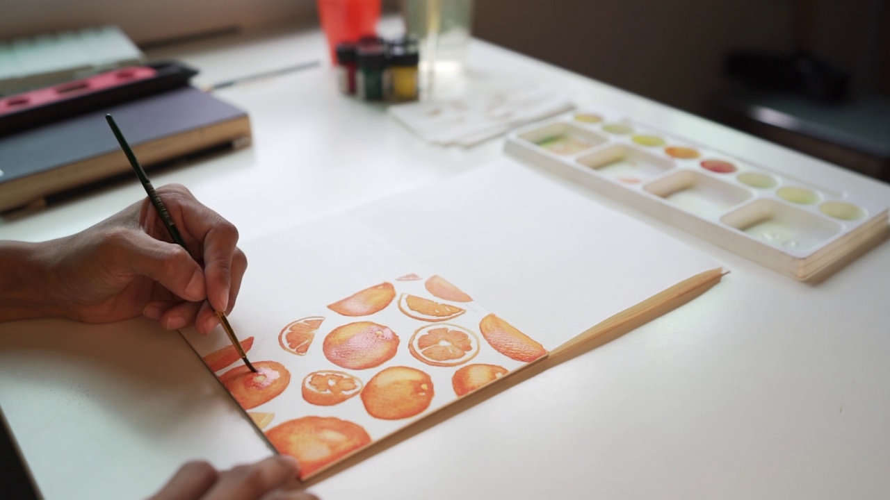 “手人”们用画笔在纸上画出果汁、橙子、橙子切片和带有橙色图案的水彩画。橙色趋势色彩概念视频下载