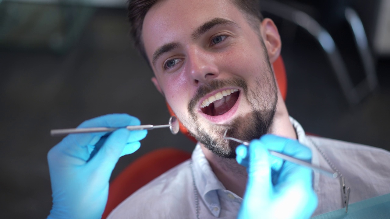 特写的面孔，自信的白人男子在牙科椅子上开放的嘴，牙医手在手套检查牙齿在慢动作。男性病人在医院接受体检。视频素材