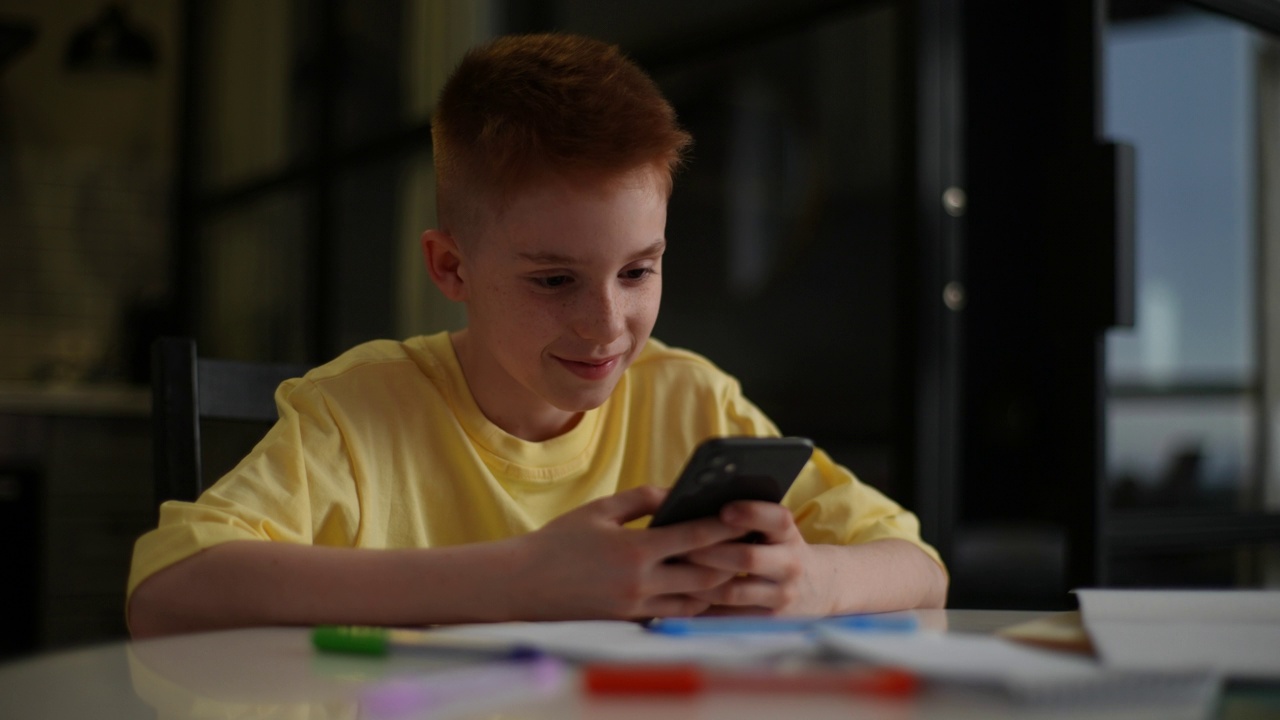 可爱的红发小男孩，穿着黄色t恤，带着雀斑坐在桌子旁，拿着智能手机，在家里玩手机应用程序，玩游戏。视频素材