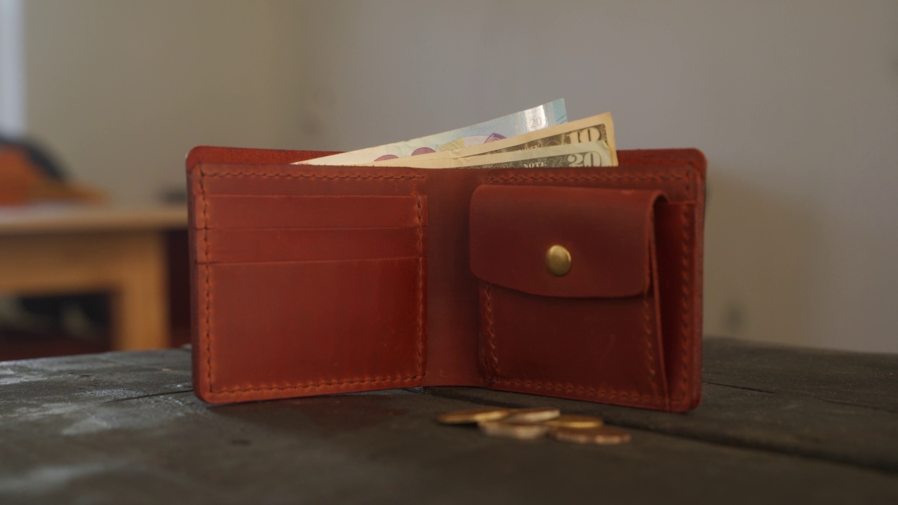 棕色双联钱包与硬币口袋和持卡人和现金是在皮革车间的深色木桌上。邮袋，货币，美元和欧元纸币，带有复制空间的硬币。财经概念。视频下载