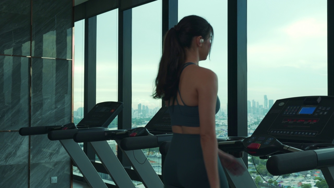 后视图的亚洲年轻的成年妇女做有氧运动步行在跑步机上，看外面看到屋顶的景色。视频素材