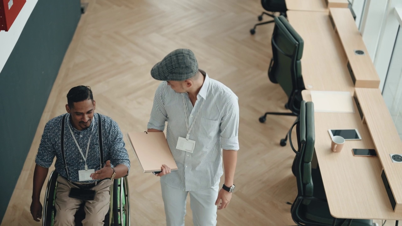 轮椅上的印度男性残疾白领与在走道走廊合作的亚裔中国男性同事讨论视频素材