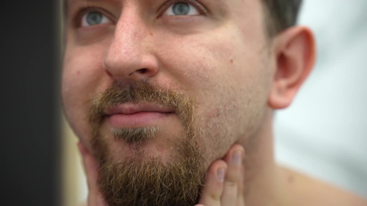 男士在剃须后将润肤油涂在脸部发炎的皮肤上，男士则在浴室用润肤油按摩脸颊和胡须。早晨日常程序，男士护肤卫生理念视频素材