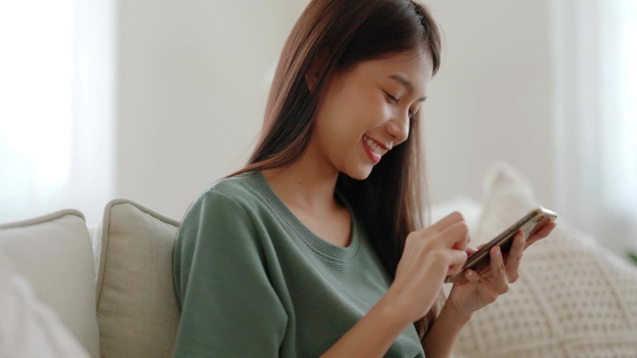 快樂的年輕亞洲女性放松在舒適的沙發上在家里用智能手機發短信，微笑的女孩使用手機聊天，瀏覽無線互聯網上的小工具，從家里網上購物視頻下載