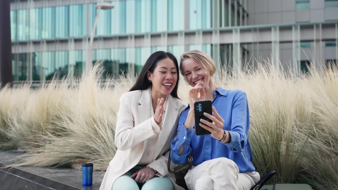 快乐的女同事坐在办公室花园用智能手机与专业的同事朋友分享视频通话视频下载