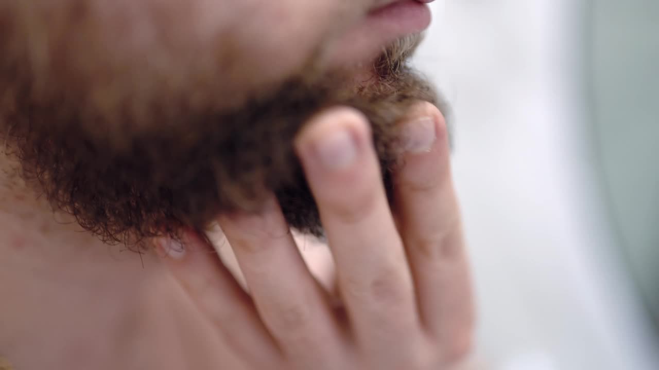 特写镜头中，男子用手摸着他浓密的胡子，男子在浴室里对着镜子做日常面部皮肤护理。针对干燥或过敏面部皮肤的问题，每日剃须，用润肤油平滑胡须和皮肤视频素材