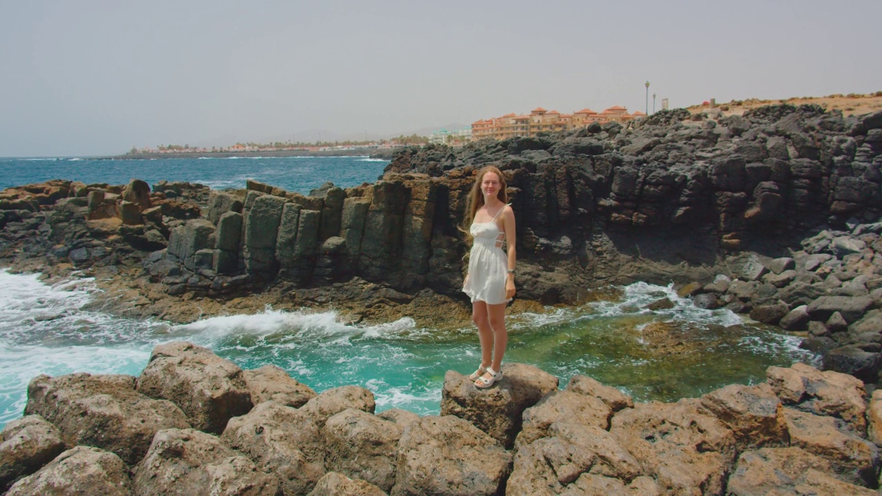 侧视图的白人妇女站在岩石上在海上享受放松阳光明媚的日子在后视图城市景观。大自然旅行海洋大浪，户外岩石火山景观，风在慢镜头。视频下载