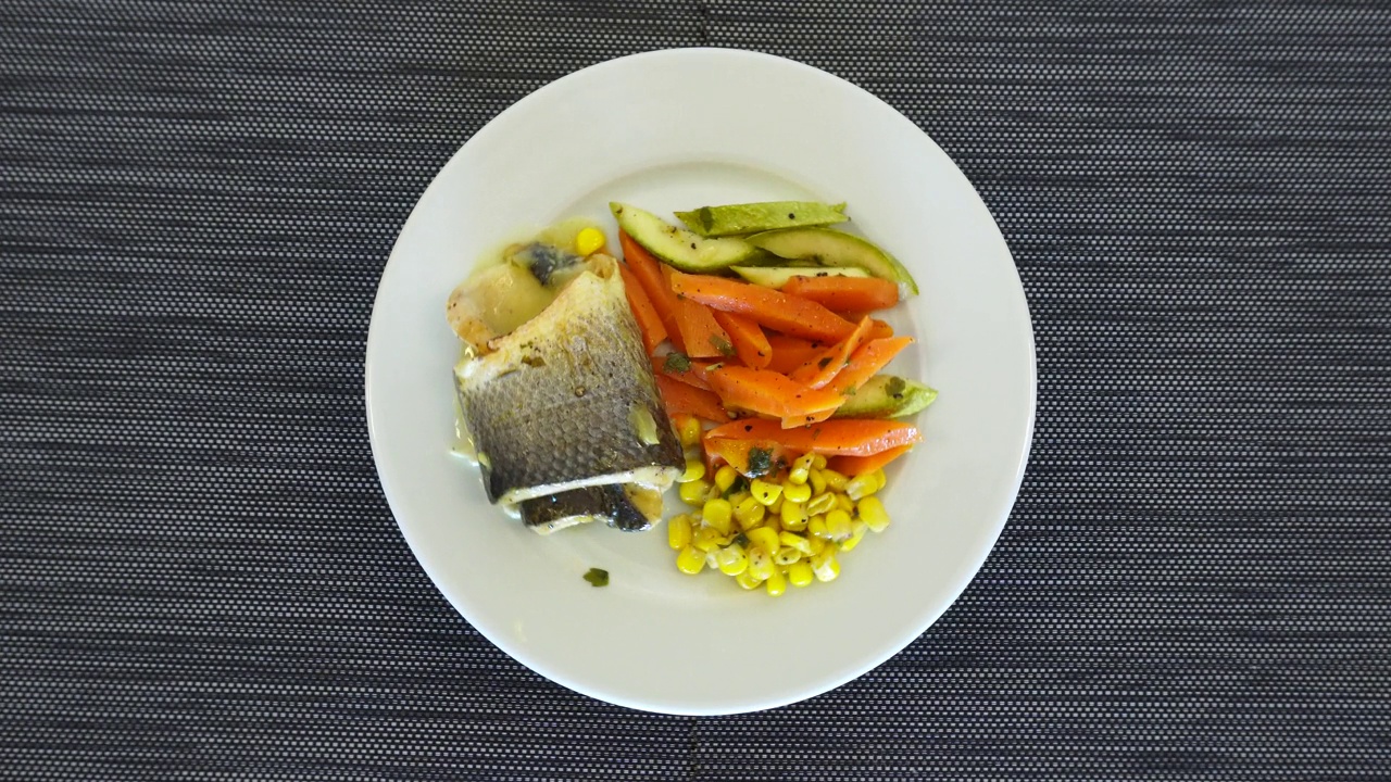 一块炸鱼片和蔬菜放在一个旋转的盘子里。胡萝卜，玉米粒和西葫芦作为配菜。从上面看。视频下载