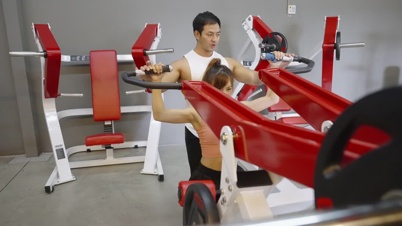 可爱的亚洲女人在举重机上专心锻炼，她的教练在健身房密切指导视频下载