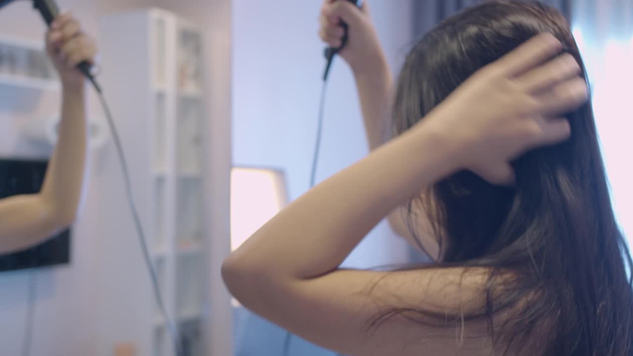 早上洗完澡后，亚洲美女拿着热吹风机在镜子前吹长黑发。女性的日常生活方式是在家照顾头发健康。视频下载