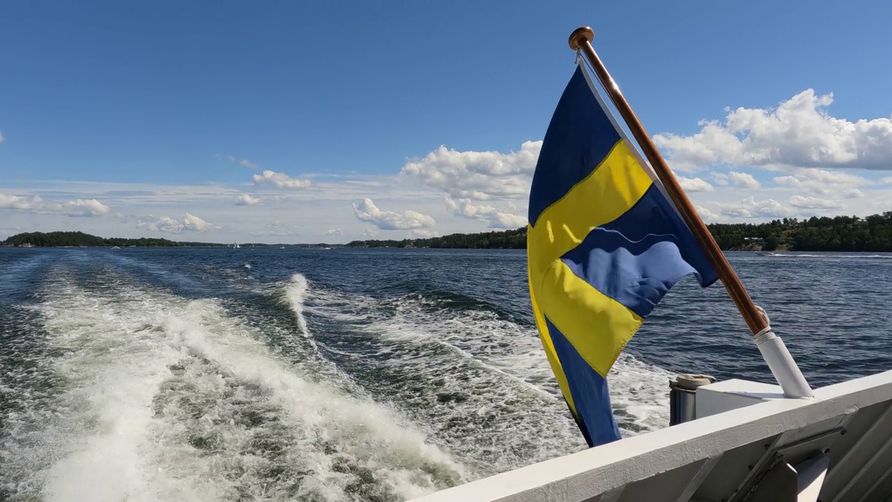 瑞典国旗在斯德哥尔摩群岛航行的船只尾部飘扬。侧面观看视频的旗帜在中间的船后甲板视频素材