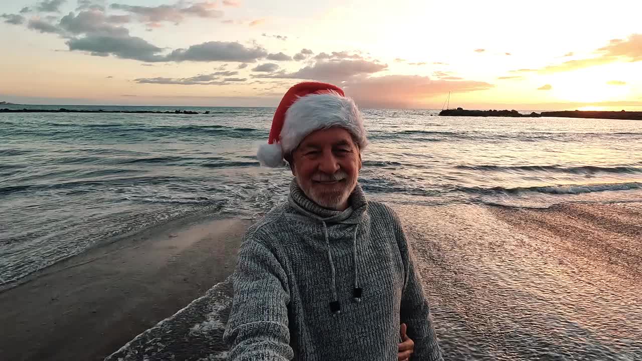 两位快乐的老人戴着圣诞帽在海滩上自拍，他们微笑着，在傍晚的日落背景下玩得很开心。一对可爱的老人看着镜头视频素材