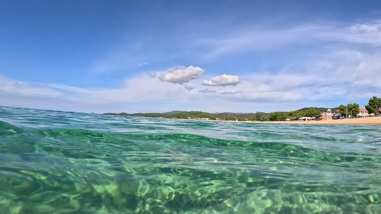 希腊哈尔基迪基岛锡托尼亚清澈的海水。半水下的沙滩底部，蓝天和云的慢动作视图。从空气和水的边界与圆顶拍摄视频素材