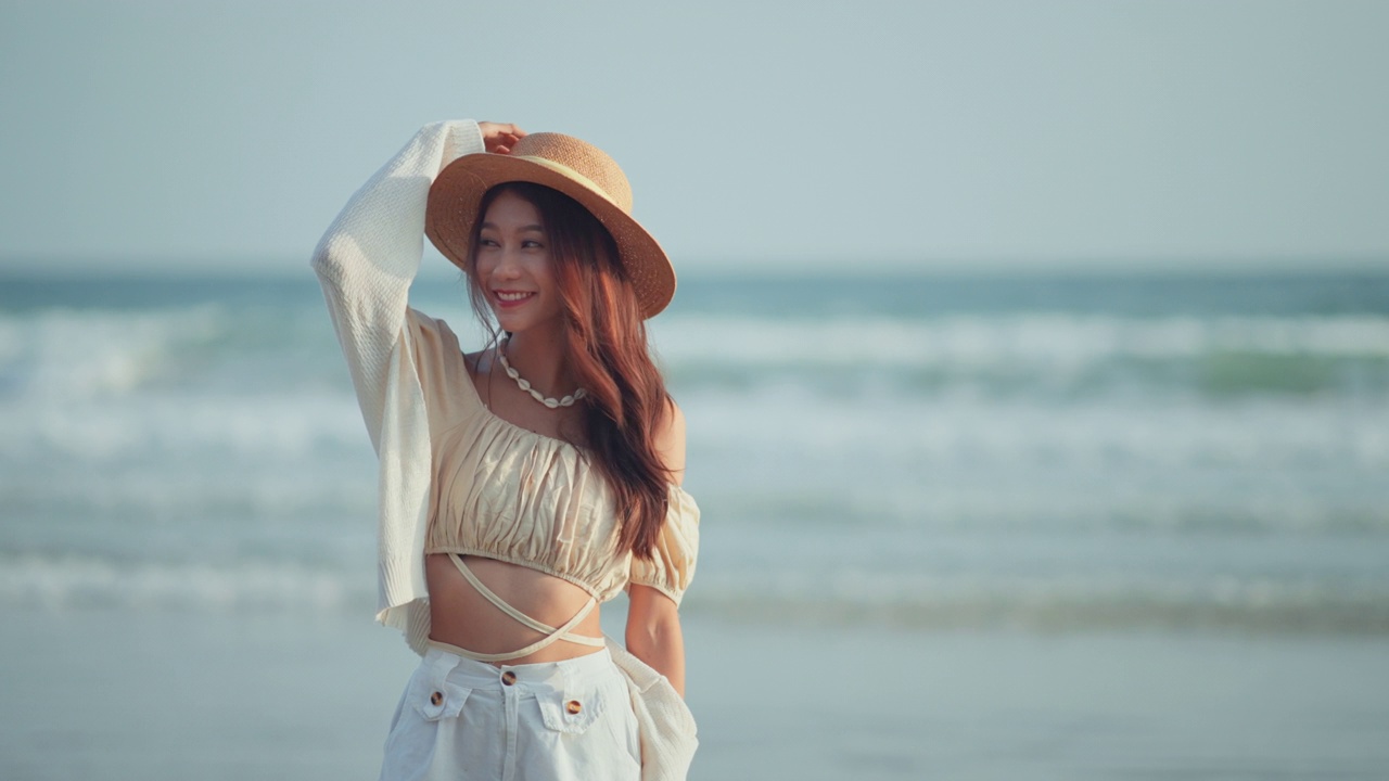 快乐的亚洲年轻女子戴着草帽漫步在热带海滩上，无忧无虑的女性微笑着享受着以海为背景的微风。旅游度假，夏日户外娱乐。视频购买