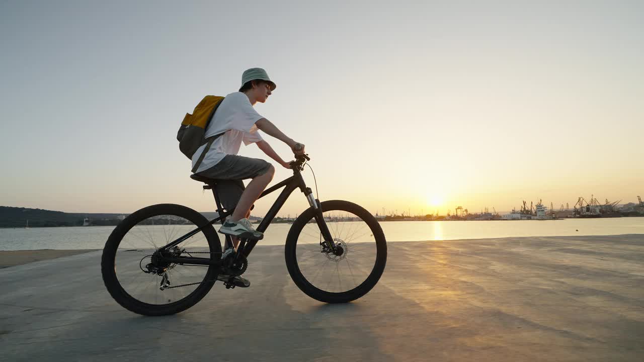 夏天炎热的日落时分，在城市港口码头的海边，少年戴着背包帽骑着自行车的剪影。一代的生活方式。自由。运动。旅行。旅游视频下载