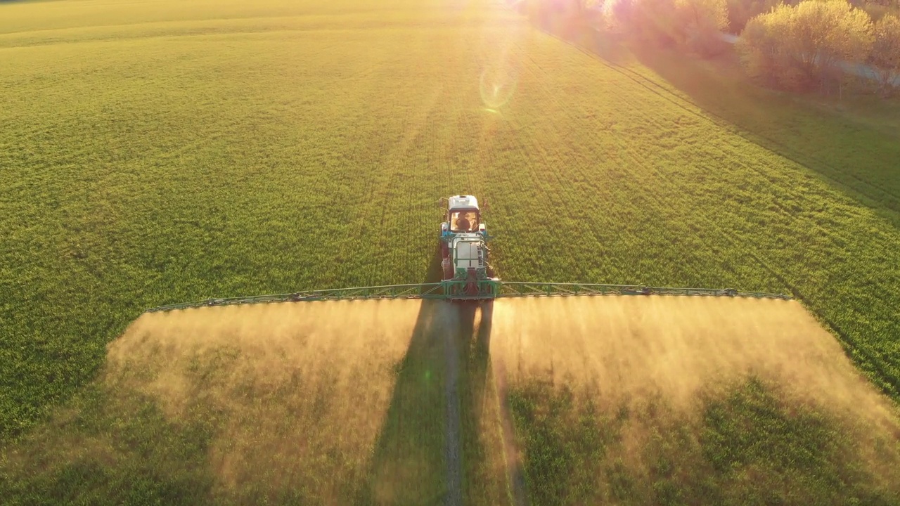 空中镜头。日落时分，农药喷雾器拖拉机在一大片绿色的田野上工作。航拍照片，旁边是一辆拖拉机在麦田里喷洒防病剂。农民喷洒大豆田。视频下载