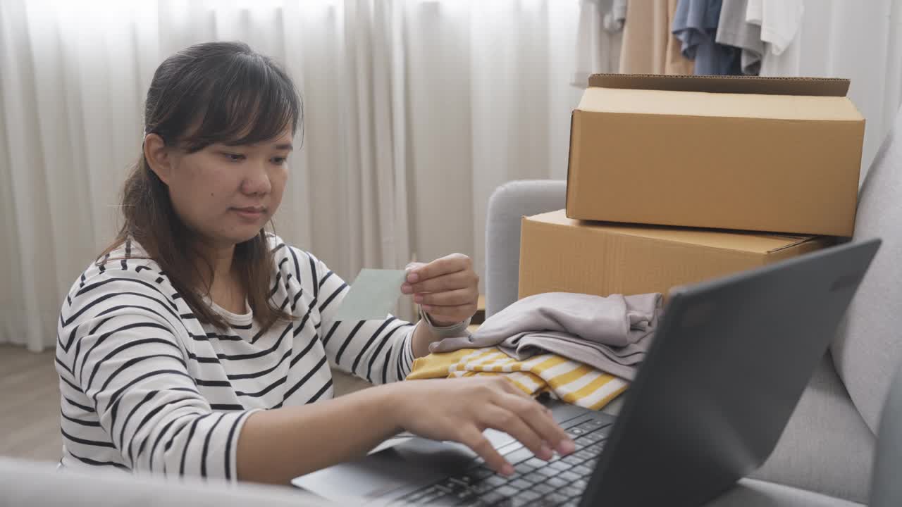 一名年轻的亚洲女子坐在地板上查看她的衣服订单数据，她的衣服是通过网购平台销售的，这是她的副业视频素材