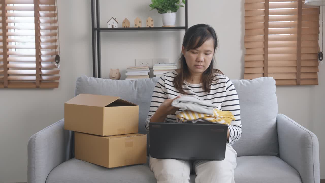 一名亚洲女子坐在沙发上，检查她在购物平台上在线销售的数据订单，然后打包发货视频素材
